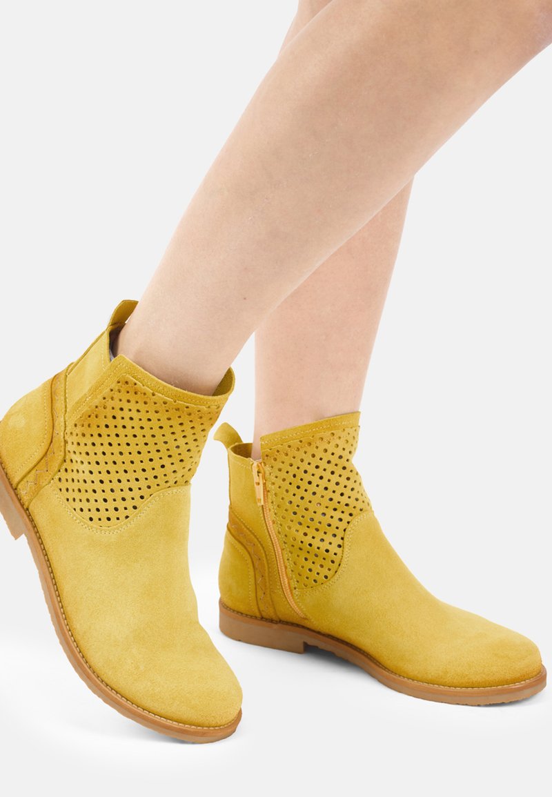 Żółte botki zamszowe z perforowanymi górnymi częściami, zamkami błyskawicznymi po bokach i niskimi brązowymi gumowymi podeszwami, noszone na bosych stopach.
