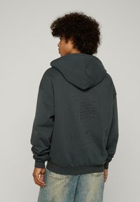Dunkelgrauer Kapuzenpullover mit lässiger Passform, weichem Material und einem erhöhten Grafikdesign eines Saftkartons auf der Rückseite mit dem Schriftzug "SELF LOVE JUICE".