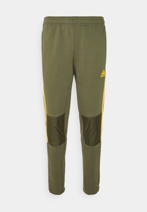 Olijfgroene Adidas trainingsbroek met gele strepen aan de zijkanten, elastische tailleband en donkerdere olijfgroene stukken op de knieën.