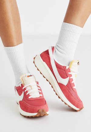 Persona que lleva calcetines blancos tobilleros y zapatillas Nike rojas con el logo blanco y suelas de goma, de pie sobre una superficie de color gris claro.