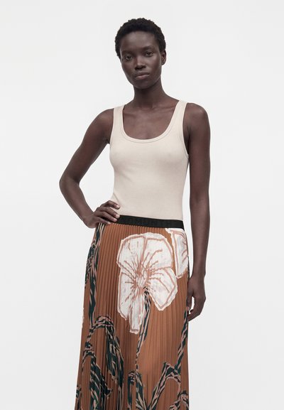 Femme aux cheveux courts et bouclés portant un haut beige sans manches et une jupe plissée marron à grand motif floral, debout la main sur la hanche.