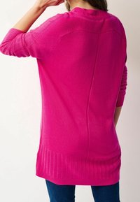 Femme portant un pull à manches longues rose vif avec un ourlet et des coutures côtelées, associé à un jean bleu foncé, vue de dos.