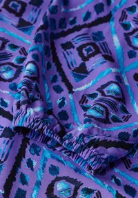 Lila Stoff mit einem geometrischen Muster und blauen sowie schwarzen Akzenten. Das Material ist dehnbar und hat geraffte Kanten für einen figurbetonten Look.