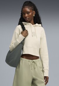 Licht beige cropped hoodie met frontlogo, gecombineerd met losse lichtgroene broek. Model houdt een zachte, donkergroene tas vast met een gekreukte strap.