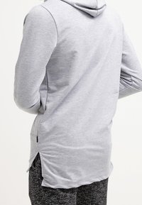 Grauer Hoodie mit einer Fronttasche, schlankem Schnitt und einem etwas längeren Rücken. Weicher Stoff mit glatter Textur, ausgestattet mit einer Kapuze und seitlichen Schlitzen.