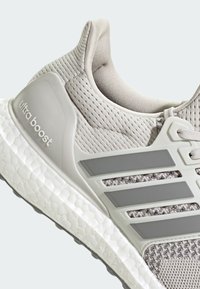 Lys grå sneakers med en teksturert mesh-overdel, som har tre fremtredende grå striper, polstret hæl og en hvit teksturert såle.