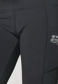 Juodos sportinės legginsai, pagaminti iš tamprios medžiagos, su šonine kišene ir baltu grafinio logotipo abstrakčiu dizainu.
