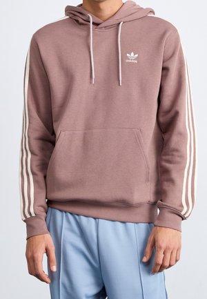 Mand iført støvet lyserød Adidas hættetrøje med hvide striber på ærmerne og lyseblå bukser, står med hænderne afslappet ned langs siderne.