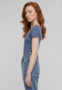 Modra kratka majica s kratkimi rokavi iz mehke bombažne tkanine, nosena s hlačami iz rahlo obrabljenega denima z visokim pasom, prikazana s strani.