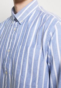 Zbliżenie na osobę ubraną w niebiesko-białą koszulę w pionowe paski z zapięciem na guziki przy kołnierzyku i kieszonką na piersi.