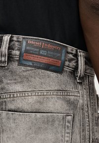 Κοντινό πλάνο ξεθωριασμένου γκρι τζιν με τμήμα επωνυμίας Diesel Industry πάνω από την πίσω τσέπη, φορεμένο από άτομο με μαύρο πουκάμισο.