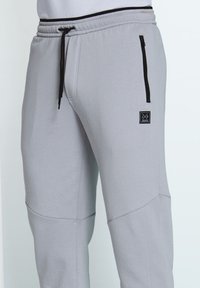 Hellgraue Jogginghosen mit einem Kordelzugbund, Reißverschluss-Taschen und strukturiertem Stoff. Mit einem kleinen schwarzen Logo-Patch am Oberschenkel.