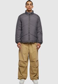 Grå pufferjakke med høy krage, glidelås foran og chevron-søm. Kombinert med beige, oversized cargo-bukser og hvite sneakers.