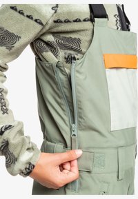 Groene geïsoleerde overall met een oranje zak, ritssluiting, verstelbare banden en zachte, gebreide fleece eronder. Gestructureerde stof.