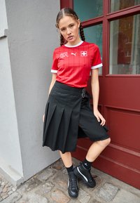 Sarkana Puma futbola krekls ar baltajiem akcentiem, apvienots ar melnu plisēto svārku ar sānu šķēlumu un melnām augstajiem sporta apaviem.