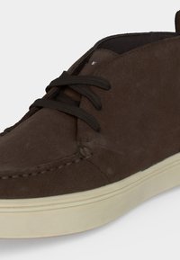 Chaussure décontractée en daim marron foncé avec lacets noirs et semelle en caoutchouc blanc cassé, vue de profil à l'avant.