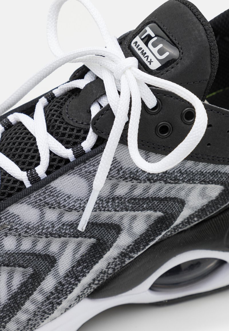 Zapatilla en blanco y negro con parte superior de malla, patrones texturizados y cordones prominentes. Cuenta con una suela de goma y un logotipo de marca visible.