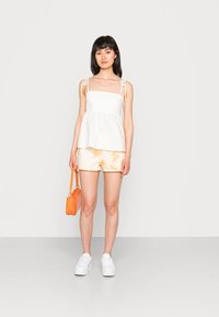 Top peplum branco com alças ajustáveis, combinado com calções em tie-dye pastel. A modelo segura uma pequena bolsa laranja, vestindo ténis brancos.