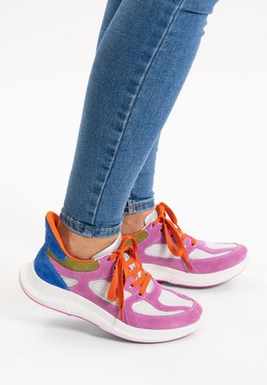 Voeten met roze, witte en blauwe sneakers met oranje veters, gecombineerd met blauwe spijkerbroeken, tegen een witte achtergrond.