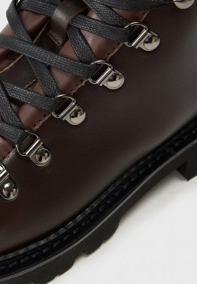 Stivale da trekking in pelle marrone con lacci neri, occhielli metallici e suola nera resistente con un evidente battistrada.