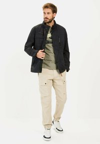 Schwarze Außensjacke mit hohem Kragen, mit Reißverschlusstaschen, getragen über einem grünen T-Shirt. Beigefarbene Cargo-Hosen mit elastischen Bündchen und Taschen. Weiße Sneaker.