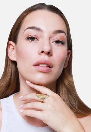 Junge Frau mit leichtem Make-up und langen Haaren, die ihr Kinn berührt, trägt einen großen goldenen Statement-Ring und ein weißes ärmelloses Top.