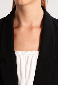 Blazer noir avec un col structuré, associé à un haut blanc loose avec un encolure élastique. Tissu lisse avec une coupe ajustée.