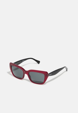 Occhiali da sole in plastica rossa con montatura quadrata e lenti grigie. Presentano braccia nere con branding bianco sull'esterno. Texture liscia.