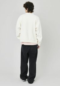 Sweat-shirt à col rond surdimensionné en blanc cassé, avec une finition texturée et un ourlet ajusté, superposé sur un haut rose. Porté avec un jean large noir.