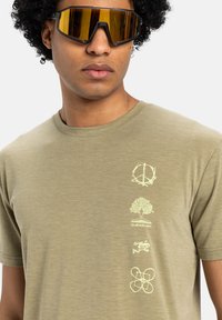 Oliwkowy t-shirt z krótkim rękawem z delikatnymi grafikami: symbol pokoju, drzewo i abstrakcyjne figury, o miękkiej, gładkiej teksturze.