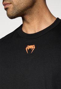 Μαύρο βαμβακερό μπλουζάκι με πορτοκαλί λογότυπο φιδιού στο στήθος. Στρογγυλή λαιμόκοψη και κοντά μανίκια με ομαλή υφή.