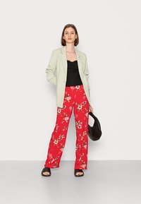 Blazer verde claro sobre uma blusa preta, combinado com calças largas vermelhas com padrão floral e sandálias pretas, segurando uma bolsa preta em textura.