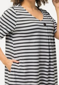 Pasiasta tunika w czarno-szarym materiale, z dekoltem w kształcie V, krótkimi rękawami, jednym guzikiem i bocznymi kieszeniami.