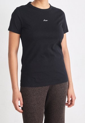 Kobieta ubrana w jednolity czarny t-shirt z krótkim rękawem z małym białym logo przy kołnierzu oraz brązowe prążkowane spodnie dzianinowe.