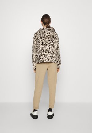 Vrouw met vastgebonden haar die een beige hoodie met stippen, beige joggingsbroek, zwarte sokken en witte sneakers draagt, staand met het gezicht naar een witte muur.