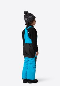 Blue and black snow overall with adjustable straps, worn over a black long-sleeve shirt, completed with a knitted black hat with a pom-pom.