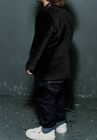 Blazer in velluto nero con due bottoni, jeans blu scuro con orlo risvoltato e sneaker bianche. Vista posteriore su uno sfondo neutro.