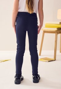 Tumši zili legingi ar gludu, pieguļošu dizainu. Aprīkoti ar aizmugurējo kabatu un vidēji augstu jostu, kopā ar spīdīgām melnām kurpēm.