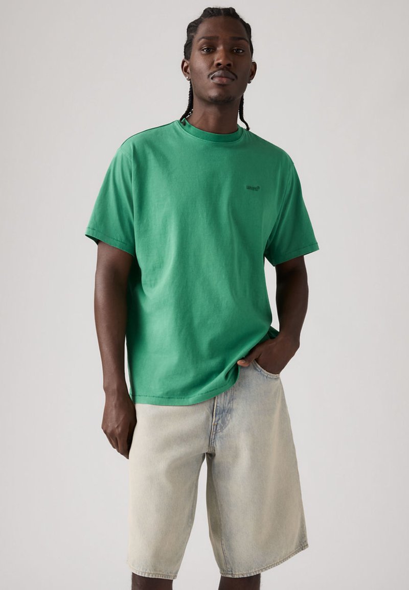Jeune homme aux cheveux tressés portant un T-shirt vert et un short en jean clair, debout avec une main dans la poche devant un fond uni.