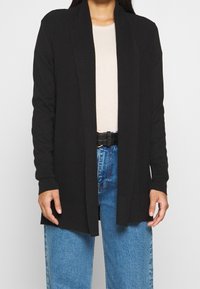 Schwarzer Cardigan mit langen Ärmeln, offener Front, gerippter Textur und seitlichen Schlitzen. Getragen über einem beigen Oberteil mit blauen hochgeschnittenen Jeans.