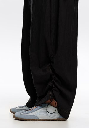 Persona con pantalones negros sueltos con cordones ajustables, sobre zapatillas azul claro con cordones y suelas de goma, sobre un suelo blanco.