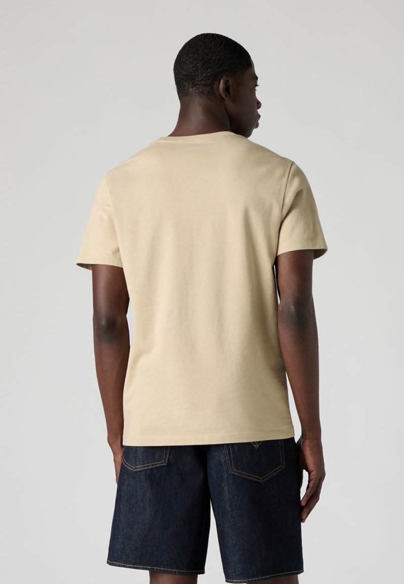 Beiges Baumwoll-T-Shirt mit Rundhalsausschnitt und kurzen Ärmeln, getragen zu dunklen Jeansshorts, mit minimalistischem Design und entspannter Passform.