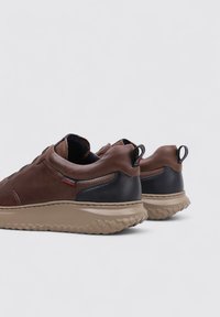 Zapatillas de cuero marrón con detalles en negro, suela de goma texturizada y tira en la parte trasera. Presentan un diseño minimalista y una silueta casual.