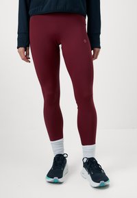 Persona vistiendo leggings marrones, calcetines blancos, zapatillas de correr en blanco y negro, y una camiseta de manga larga oscura contra un fondo liso.