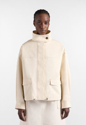 Mujer con cabello trenzado que lleva una chaqueta color crema de cuello alto con bolsillos de solapa y falda a juego sobre un fondo sencillo.