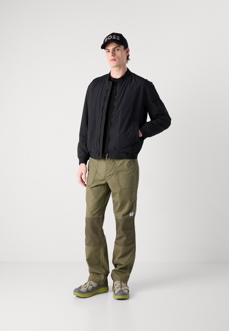 Czarna kurtka bomberka z obszytymi mankietami, oliwkowe spodnie cargo z łatkami na kolanach, szare i zielone sneakersy oraz czapka na czarno z logo "BOSS".