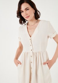 Cream-colored linen dress with short sleeves, V-neck, and button front. Features a gathered waist, two side pockets, and a relaxed fit.