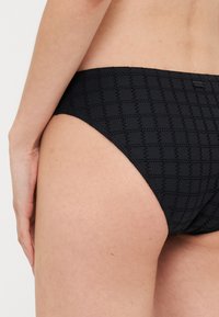 Svart bikiniunderdel med en texturerad, geometrisk mönstring. Designen är låg midja med en slät finish och minimalistisk stil.