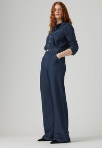 Chemise en denim avec col et boutons à l'avant, associée à un jean large haute taille bleu foncé, doté de poches latérales. Chaussures noires pointues.