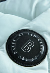 Svijetloplava podstava s tablicom od crnog platna u obliku kruga. Tablica sadrži bijeli tekst i logo "B" za Ted Baker. Glatka tekstura.